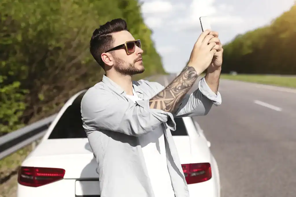 A man with sunglasses taking a picture of a white car on the roadside, highlighting auto detailing service branding.